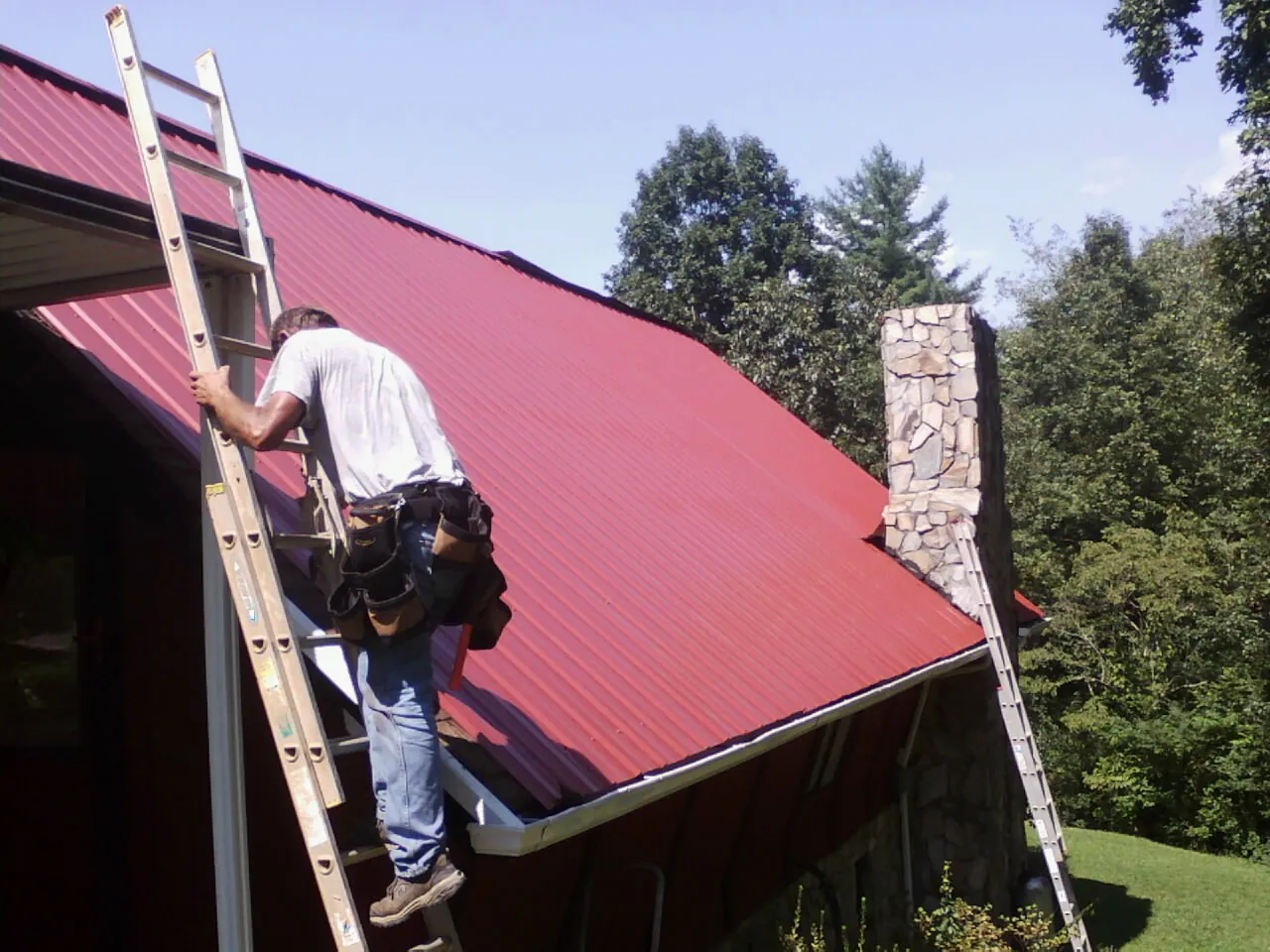 Roofer on a ladder beside a red metal roof and stone chimney — Soffit & Fascia Repair by Roofing Brothers in Rock Tavern